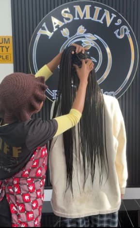 Stylist working on long dark braids in front of the Yasmin's Beauty Salon sign